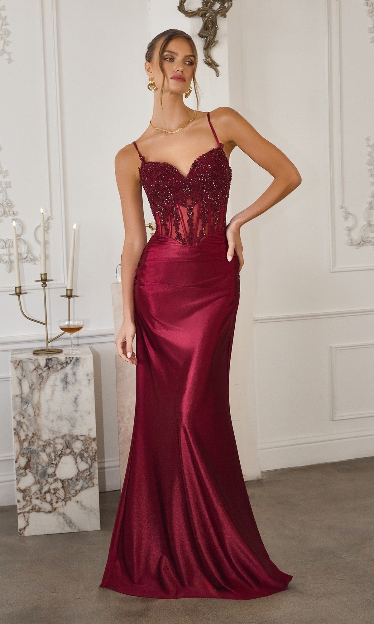 A woman in a fitted Long Prom Dress: Amoris T819 in burgundy, featuring spaghetti straps and an embellished corset bodice, stands indoors by a marble pedestal with candles.
